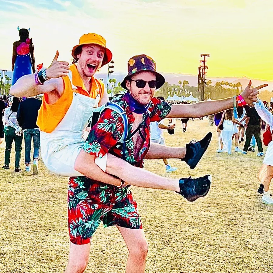 Two friends in bright outfits at Coachella 2022 at sunset, one lifted onto the other's back