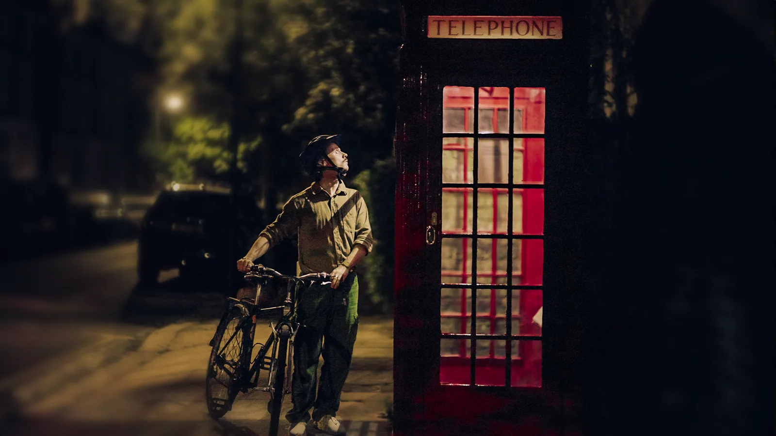 Still from RECALL: a man in a cycling helmet, holding his bike, looks up at a glowing red telephone box on a London street at night